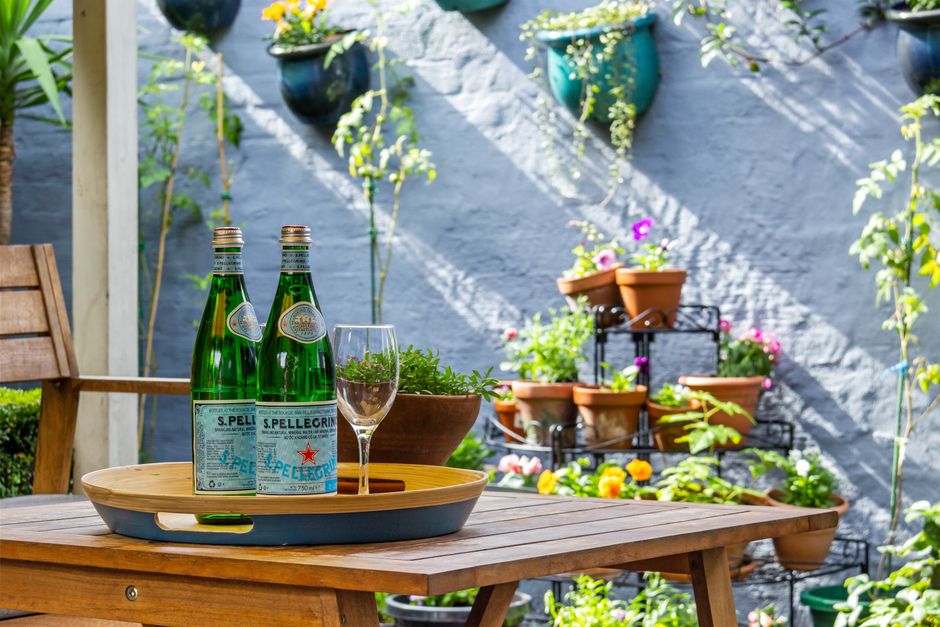 A small garden patio featuring a wooden table with a black tray holding two green bottles of S. Pellegrino sparkling water, an empty wine glass, and a plant in a terracotta pot. The background displays a gray textured wall with several terracotta pots and colorful flowering plants arranged on a black metal tiered plant stand. To the left, a wooden garden chair is partially visible. The garden area is decorated with cascading plants and leaves, with sunlight casting shadows on the wall, creating a bright and inviting outdoor space suitable for garden maintenance or leisure activities. Gardeners Richmond offers professional gardening services to maintain such vibrant and well-organized garden layouts, including hedge trimming, lawn care, and outdoor landscaping.